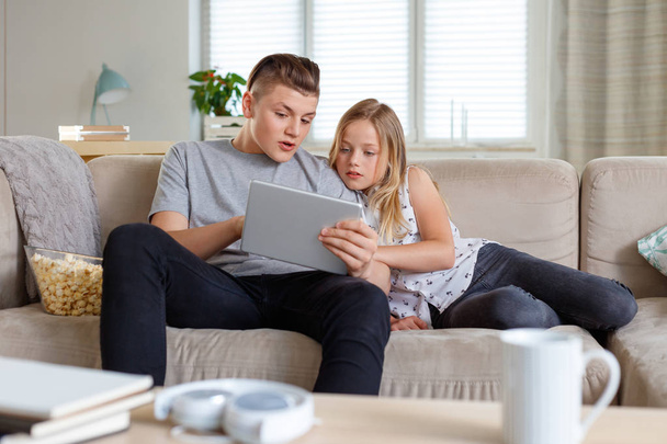 Brother and sister sitting on sofa at home in the living room and using digital tablet together. - 写真・画像