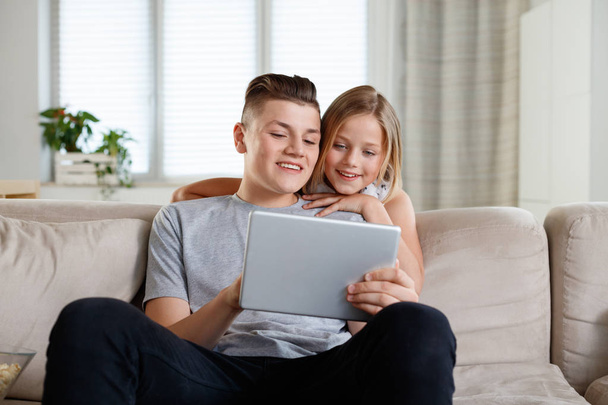 Brother and sister sitting on sofa at home in the living room and using digital tablet together. - Foto, Imagen