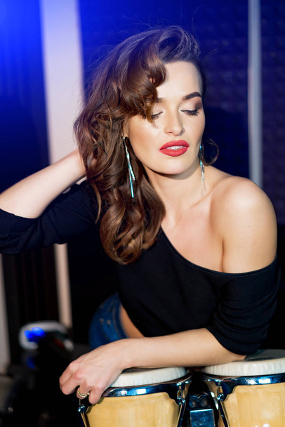Attractive model with wavy curls, red lips and long earings leans on the drums in music studio. Portrait of sexy woman posing with closed eyes. - Foto, afbeelding