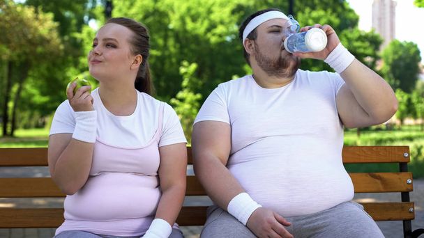Fat couple on bench, man drinking water, woman eating apple, healthy lifestyle - Foto, immagini