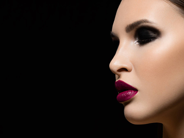 Close-up of the beauty of a woman's face with evening make-up emphasizing her expressive facial features. Smoky eyes, marsala-colored lips and clear skin. On an isolated black background - Photo, image