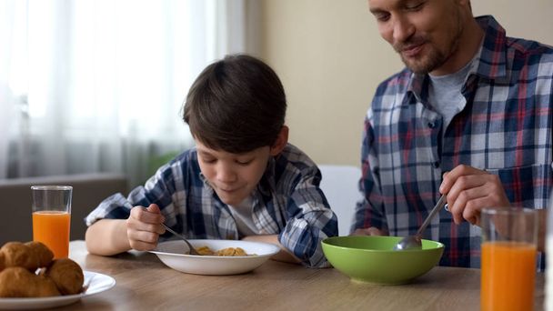 Agradable hijo y padre comiendo sabrosos copos de maíz en el desayuno, tradición de la mañana
 - Foto, Imagen