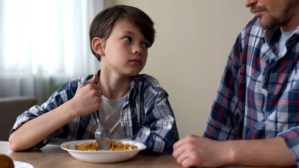 Little sad boy mixing cornflakes with spoon, looking at father, poor appetite - Foto, Imagen