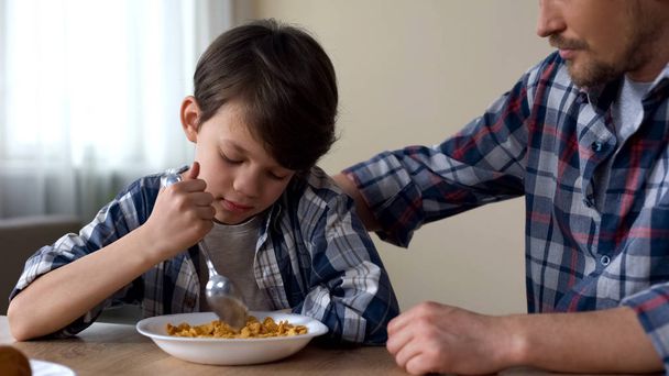Sad male kid mixing cornflakes with spoon, father stroking child, poor appetite - Фото, зображення