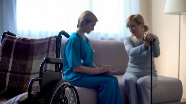 Nurse and aged woman sitting on sofa at hospital, discussing treatment, support - 写真・画像