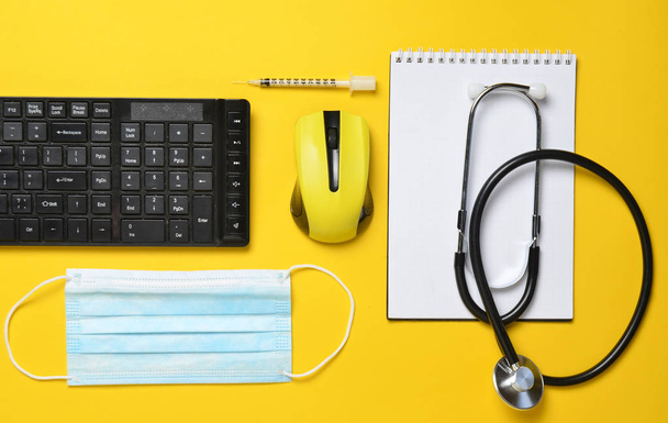Nykyaikaisen lääkärin työpaikka. Näppäimistö, langaton hiiri, muistikirja, stetoskooppi, ruisku keltaisella pohjalla, ylhäältä katsottuna, minimalistinen tren
 - Valokuva, kuva