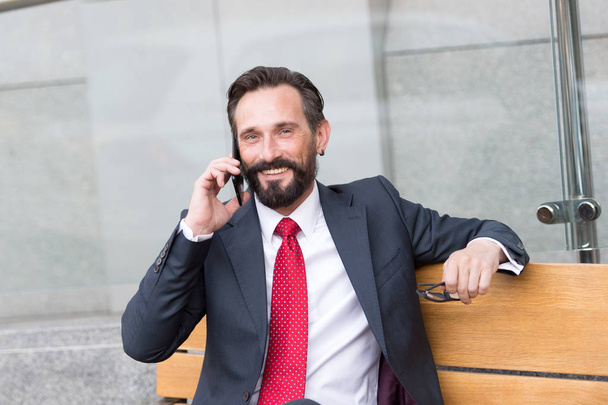 Ottime notizie. Ritratto di banchiere barbuto sorridente seduto su una panchina di legno mentre parla al telefono ed esprime ottimismo
 - Foto, immagini
