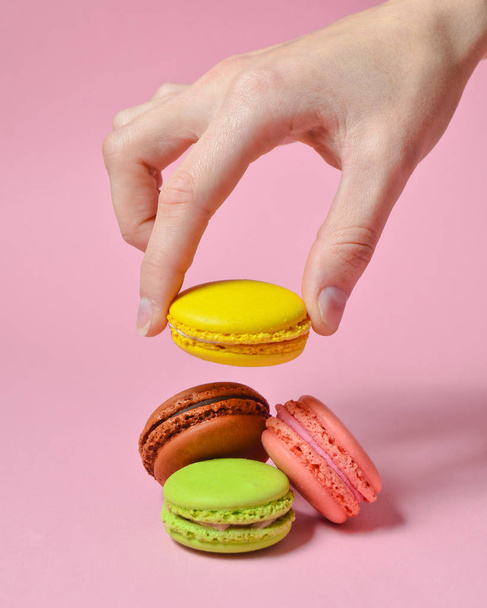 Γυναικείο χέρι χαμηλώνει το κίτρινο αμυγδαλωτά μπισκότα. Πολλοί macaroons σε ένα ροζ παστέλ φόντο. Minimalis - Φωτογραφία, εικόνα