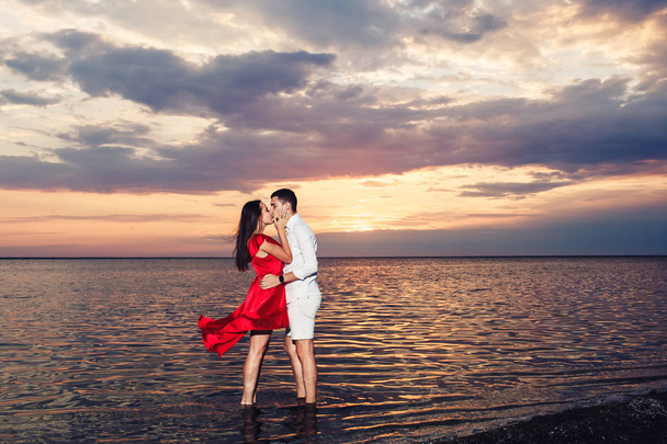 νεαρό ζευγάρι στην αγάπη φιλιά στέκεται στη θάλασσα με φόντο ένα μαγευτικό ηλιοβασίλεμα. - Φωτογραφία, εικόνα