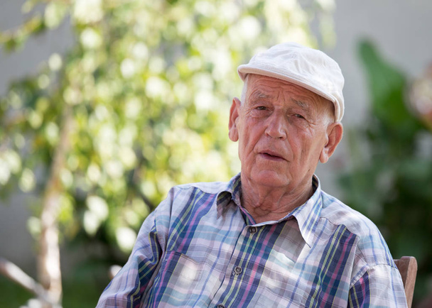 Portrait of senior man with cap sitting in park with green tree in background - Photo, Image