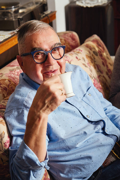 ευτυχισμένος ηλικιωμένους άνθρωπος κάθεται στον καναπέ και απολαμβάνει καφέ. - Φωτογραφία, εικόνα