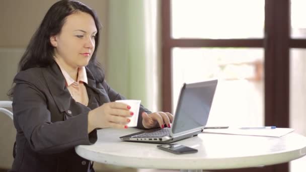Young woman at a table in a cafe, working behind a laptop and drinking coffee. Slow mo. The average plan of the camera moves from right to left. - Imágenes, Vídeo