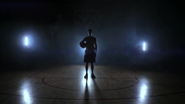 Basketbol oyuncusu karanlık bir oyun alanı üzerinde duruyor ve topu elinde tutar ve yavaş hareket ve duman çevresinde bir sırt ile karanlıkta kameraya benziyor - Video, Çekim