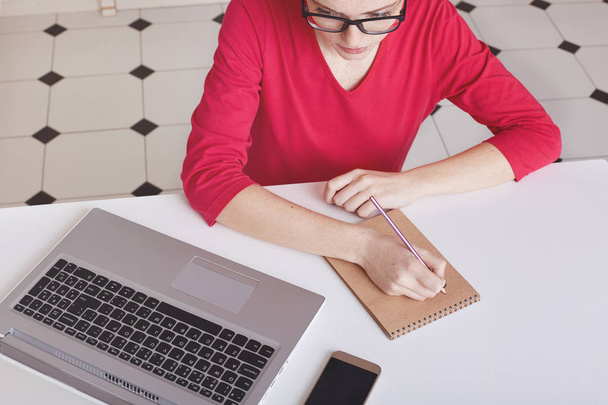 Cropped top view of busy female writer makes notes in spiral notepad, wears transparent glasses and red sweater, focused in screen of laptop, sits at white desk, rewrites information from internet - Photo, image