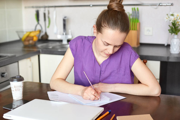 Busy female works from home, writes down something in spiral notepad, sits at kitchen table, surrounded with takeaway coffee, laptop and mobile phone, studies financial data, plans budget on next year - 写真・画像