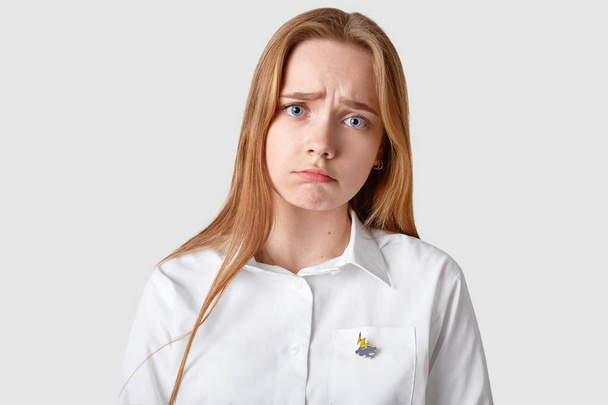 Desperate displeased sad young lady with blue eyes, long hair, purses lips, had dissatisfied facial expression, dejected by negative news, dressed in formal shirt, isolated over white background - 写真・画像