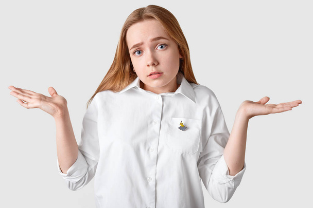 Upset clueless girl shruggs shoulders, has indignant facial expression, cant make decision, looks confusingly at camera, wears elegant shirt, isolated over white background. Unaware youngster indoor - Fotografie, Obrázek