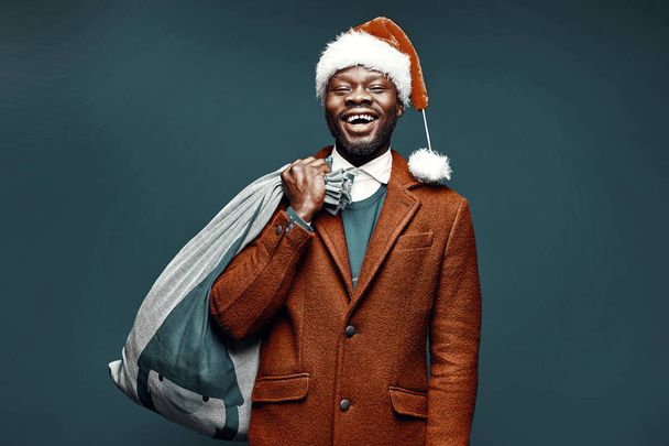 Modern Santa Claus. Smiling emotional man posing in brown coat and red sweater, with santa hat and bag. Studio shot, green background - Fotó, kép