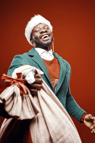 Modern Santa Claus. Smiling emotional man posing in green coat and red sweater, with santa hat and bag. Studio shot, red background - Zdjęcie, obraz