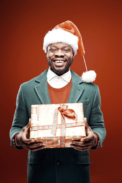Happy man in santa claus hat, green coat, with gift in hands. Studio shot, red background  - Fotografie, Obrázek