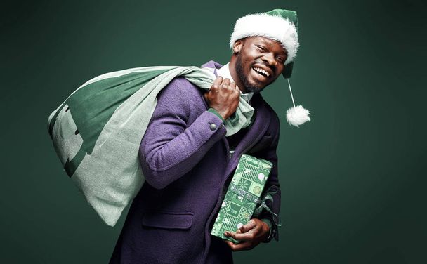 Modern Santa Claus. Smiling emotional man posing in violet coat, with green santa hat and bag and present. Studio shot, green background - Photo, Image