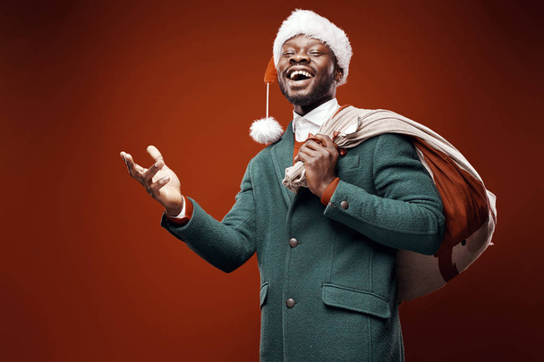 Modern Santa Claus. Smiling emotional man posing in green coat and red sweater, with santa hat and bag. Studio shot, brown background - Foto, Imagen