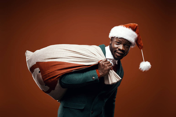 Modern Santa Claus. Confused man posing in green coat and red sweater, with santa hat and bag. Studio shot, brown background - Foto, afbeelding