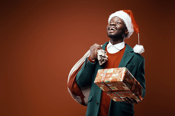 Modern Santa Claus. Smiling emotional man posing in green coat and red sweater, with santa hat and bag and present. Studio shot, brown background - Photo, image