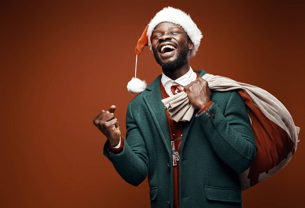 Modern Santa Claus. Smiling emotional man posing in green coat and red sweater, with santa hat and bag. Studio shot, brown background - Photo, image