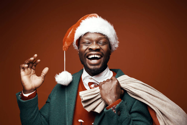Modern Santa Claus. Smiling emotional man posing in green coat and red sweater, with santa hat and bag. Studio shot, brown background - Zdjęcie, obraz