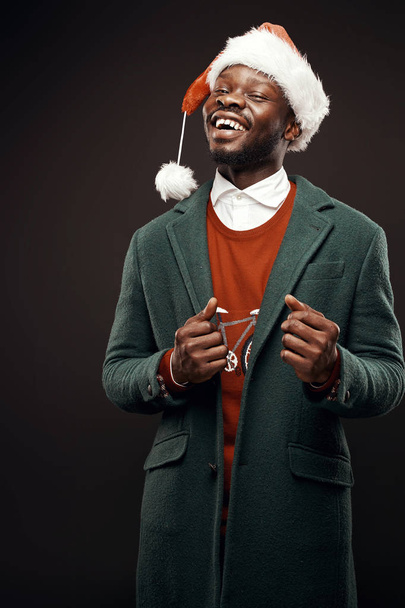 Modern Santa Claus. Smiling man in green coat and red sweater, with santa bag in hand. Studio shot, black background - Фото, зображення