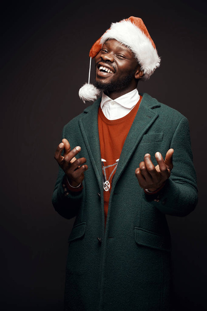 Modern Santa Claus. Smiling man in green coat and red sweater, with santa bag in hand. Studio shot, black background - Photo, image