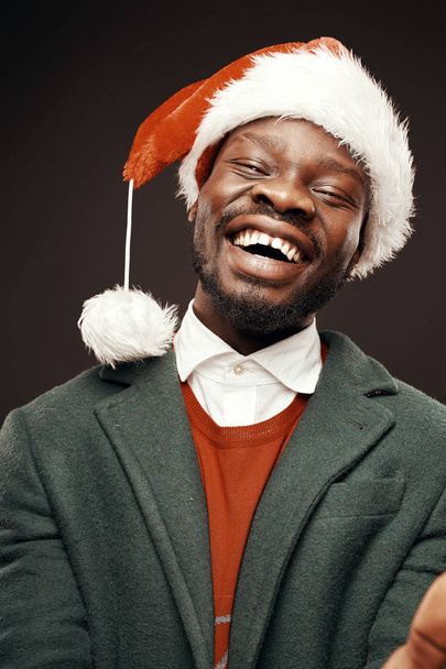 Modern Santa Claus. Smiling man in green coat and red sweater, with santa bag in hand. Studio shot, black background - Photo, Image