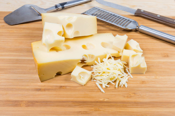 Queijo suíço parcialmente fatiado e ralado com grandes furos em uma tábua de corte de madeira de bambu contra faca de queijo, cortador e ralador
 - Foto, Imagem