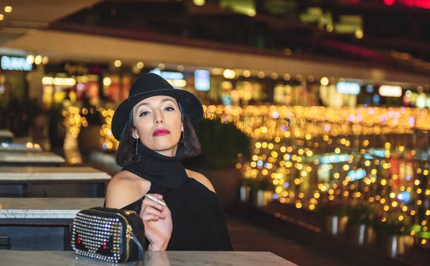 Atractiva mujer hermosa con sombrero negro y vestido se sienta por la noche con las luces de la ciudad en el fondo. - Foto, Imagen