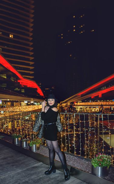 Atractiva mujer hermosa con sombrero negro y vestido posando por la noche con las luces de la ciudad en el fondo. - Foto, imagen