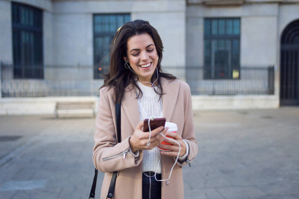 Aufnahme einer hübschen jungen Frau, die ihr Handy benutzt, während sie eine Tasse Kaffee auf der Straße hält. - Foto, Bild