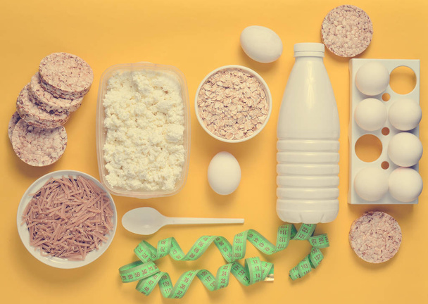 Diet, healthy food. Bottle of yogurt, crispy round bread, buckwheat noodles, oatmeal, cottage cheese, egg tray, ruler on a yellow background. The concept of losing weight. flat lay, top vie - Photo, Image