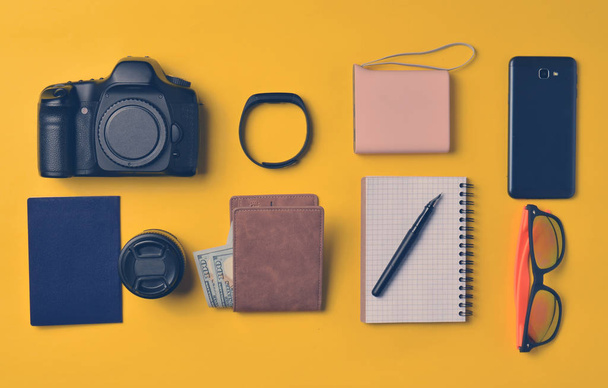 Gadgets and accessories layout on a yellow background. Power bank, photographic equipment, purse with dollars, smart clock, smartphone, notebook, sunglasses. The concept of travel, objects, top vie - Photo, Image