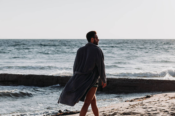 Young man in a bathrobe on the beach in sunset - Фото, изображение