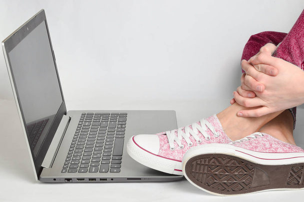 Piernas femeninas en jeans, zapatillas y un portátil sobre un fondo blanco. Espacio de trabajo freelancer. Descanso en el trabajo, falta de idea
 - Foto, imagen