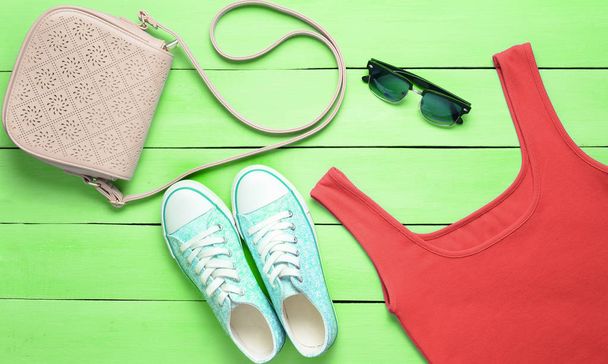 Κόκκινο T-shirt, πάνινα παπούτσια, δερμάτινη τσάντα, γυαλιά ηλίου σε ένα μπλε ξύλινο τραπέζι. Γυναικεία ενδύματα, υποδήματα και αξεσουάρ, κάτοψη, επίπεδη Λα - Φωτογραφία, εικόνα