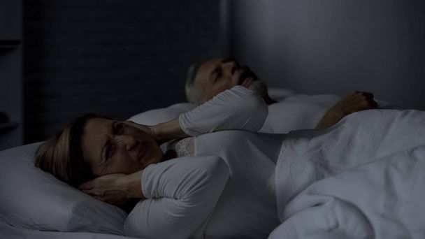 Aged lady pressing hands against ears in bed due to husband snoring loudly - Photo, image