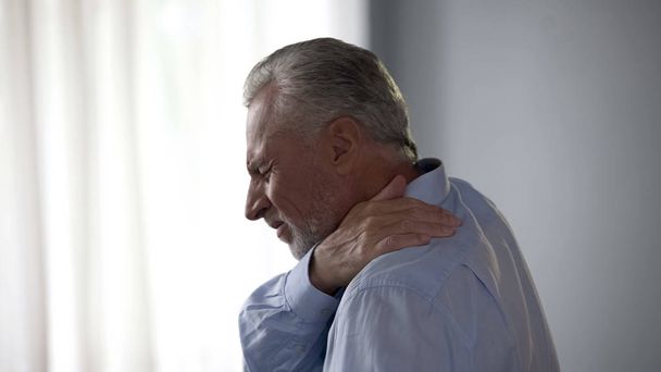 Hombre mayor tocando el cuello en dolor agudo de pie de lado, dolor en la columna vertebral, salud
 - Foto, Imagen