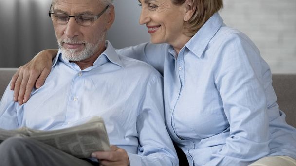 Retiree husband reading newspaper on sofa, woman beside hugging him, love - Foto, Imagen