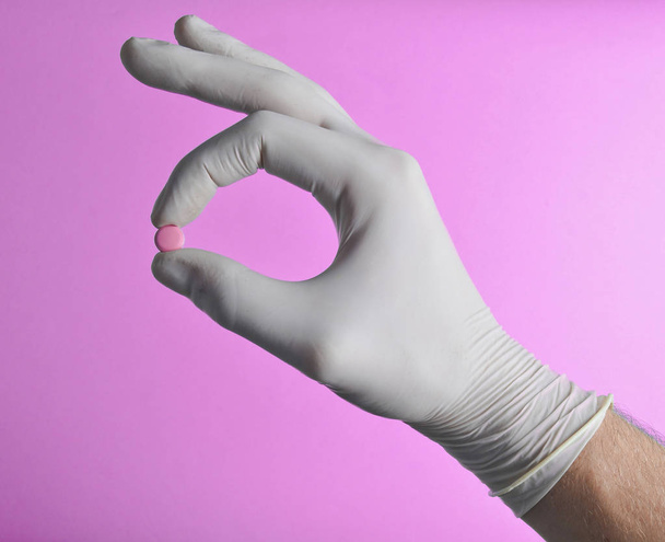 Die Hand eines Sanitäters mit weißen Latexhandschuhen hält eine rosa Tablette auf blauem Hintergrund. medizinisches Konzept - Foto, Bild