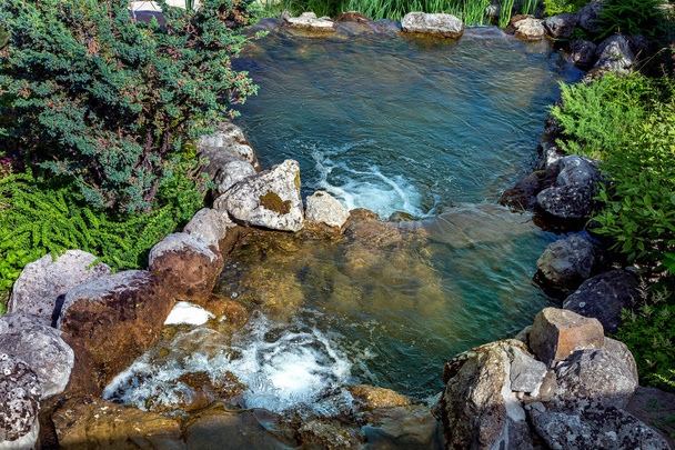 Cascata cascata artificial forrada com pedras em torno do fluxo de água ao redor da lagoa plantas plantadas
. - Foto, Imagem
