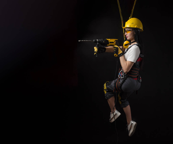 brunette girl in special clothes and a worker in a helmet posing on a black background with a working tool - Photo, Image