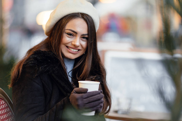Jolie femme debout dans la rue par temps nuageux avec une tasse de café chaud, buvez-le et souriez à la caméra tout en parlant au téléphone avec un fond d'espace de copie. - Image
 - Photo, image