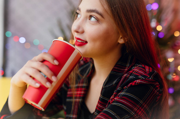 Nainen juo kuumaa kahvia istuessaan kahvilassa. Muotokuva tyylikäs hymyilevä nainen talvivaatteissa juominen kuumaa kahvia. Talvinen naistyyli. - Kuva
 - Valokuva, kuva